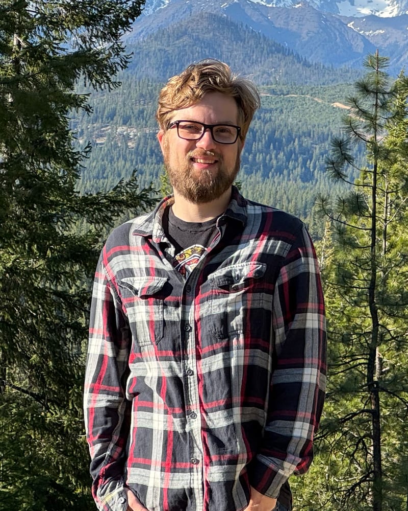 Ryan, Tradely's founder, in a plaid flannel shirt in front of pine trees and mountains